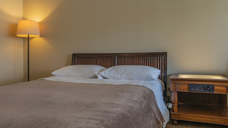 The bed is covered with white sheets and a beige terry-cloth bedspread. Two pillows at the headboard. Wooden bedside table. The floor lamp in the corner is lit. A bedroomの写真素材