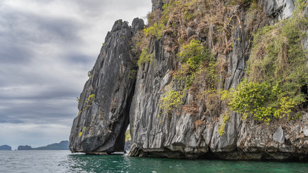 A picturesque cliff in the ocean. There is green tropical vegetation on the steep, furrowed slopes. The gap between the rocks is visible. Clouds in the sky. Philippines. Palawanの写真素材