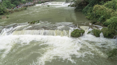 A picturesque mountain river. The water foams on the rapids. Green vegetation on boulders in the riverbed. Tourists are walking along the embankment. China. Huangguoshu Waterfall.の写真素材