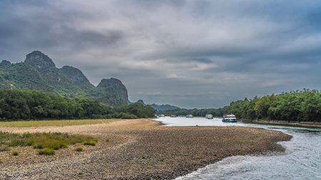 Tourist sail ships along the picturesque river. The riverbed bends. Foam on the pebbly shores. Lush green vegetation. Mountains against the sky and clouds. China. Lee River.の写真素材