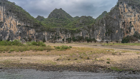 The pebbly shore of a picturesque river. Green grass, shallow pools. Coastal mountains with steep slopes and green vegetation. Clouds in the sky. China.の写真素材