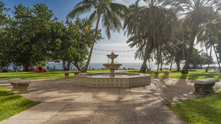 Beautiful two-tiered fountain in the park. Stone benches all around. Green grass on lawns. Palm trees against a background of blue sky and clouds. The ocean is far away.の写真素材