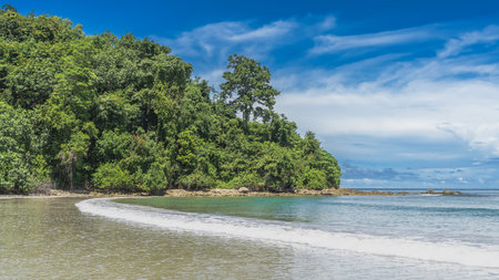 The waves of the turquoise ocean foam and spread over the sandy beach. A hill with green tropical vegetation against a background of blue sky and clouds. Malaysia. Borneo.の写真素材