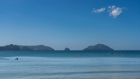 Beautiful seascape. A calm turquoise ocean, silhouettes of people in the water, orange buoys, a boat in the distance. Mountains on the horizon, against a blue sky, clouds.の写真素材