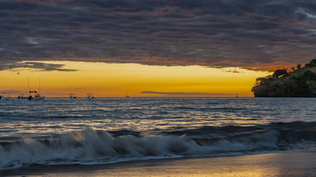 A bright sunset on a tropical island. Ocean waves are foaming on a sandy beach. Clouds are highlighted in pink. The sky is orange on the horizon. Silhouettes of boatsの写真素材