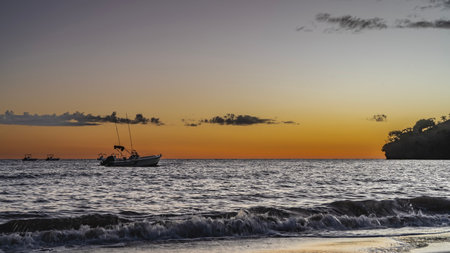 An evening after sunset in the tropics. Ocean waves are foaming on a sandy beach. Silhouette of a boat in the sea. The sky is highlighted in orange near the horizon. The clouds.の写真素材