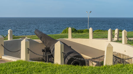 An old historical cannon on the shore of the bay. The barrel of the old cannon is pointed at the ocean. The fence is made of concrete pillars and chains.の写真素材