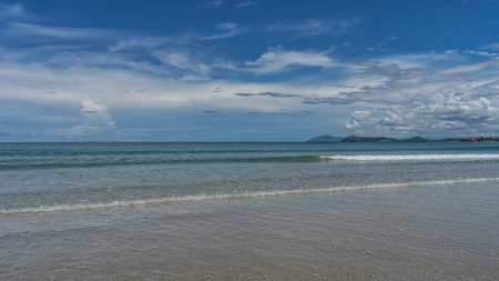 The long waves of the turquoise ocean are calmly rolling towards the shore, foaming, spreading across the sandy beach. Mountains on the horizon. Blue sky, clouds. Malaysia. Borneo.の写真素材