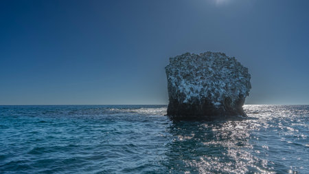 A small rocky island in the ocean. There are traces on the slopes. The waves are beating against the cliff, foaming. Glare on the water. Clear blue sky.の写真素材