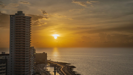 Sunset over the ocean. The sun is low. The orange sky. A sunny path on the water. Malecon embankment along the coast. In the foreground is a multi-storey city building. Cuba.の写真素材