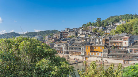 The ancient Chinese city of Fenghuang. Old buildings are closely built on the mountainside. People are walking along the river embankment. Green vegetation in the foreground.の写真素材