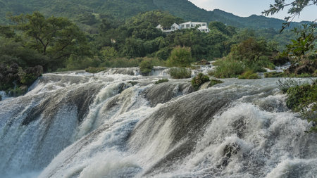 A stormy mountain river. Powerful streams of water foam on rapids and boulders. Splashes. Close-up. Green vegetation on the shore. A white building on a hill. China. Silver Chainの写真素材