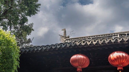 Bright red traditional Chinese lanterns are suspended from the edge of the roof. Carved cornice, the figure of a dragon's head. Green tree branches against the sky and clouds.の写真素材