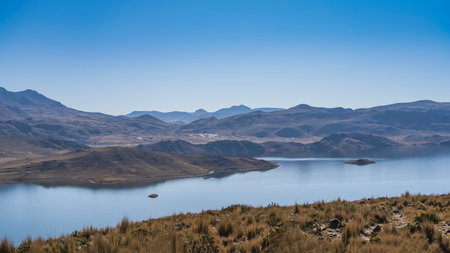 The beautiful calm alpine lake is surrounded by hills. Dry yellow grass on the shore. Clear blue...
