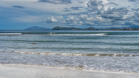 Beautiful seascape. A sunny day. The waves of the turquoise ocean roll towards the shore, foam, and spread across the beach. Mountains on the horizon. Blue sky, clouds.の写真素材