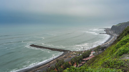 View of the ocean and the embankment from a height. The waves are rolling towards the shore, foaming. Cars drive along the highway on the shore. Green vegetation on the hillsides.の写真素材