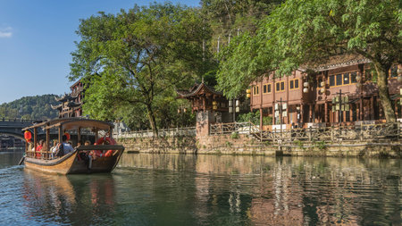 A tourist wooden boat is sailing along a picturesque river. There are traditional Chinese buildings and green vegetation on the shore. The fence along the embankment. Reflectionの写真素材