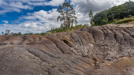 Colorful sand hill on a background of blue sky and clouds. Furrows, scattered stones on the slope. Green vegetation. Mauritius.の写真素材
