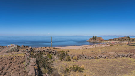 There is a sandy beach with sun umbrellas on the shore of the lake. Boulders, dry grass on the hillside. Clear blue sky. Copy space. Peru. Lake Titicaca. Taquile islandの写真素材