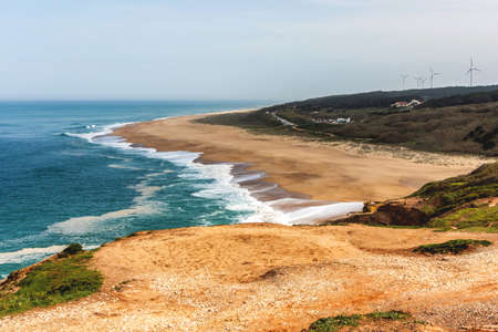 A beautiful landscape of the burgundy Atlantic Ocean and the sandy yellow shore with white foam from the waves. In the background are several houses and wind turbines of energy. Portugal, Nazare.の写真素材