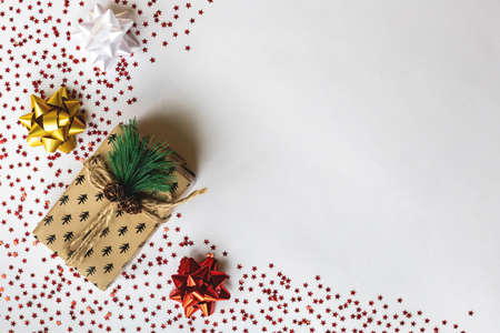 Christmas gift in brown paper tied with a scourge with cones and a branch of a Christmas tree, near bows of different colors near the gift on small stars on a white background, place for text, flatlayの写真素材