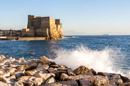 Seascape against the background of Castel dell'Ovo with a coastline of large light stones, a splash of snow-white water in the bright sun, mountains on the horizon and a ship in Naples, Italyの素材 [FY310178832793]