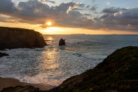 Portuguese sunset landscape with the suns rays through the clouds on the shores of the Atlantic Ocean with sheer high cliffs and foamy waves. A small island is visible on the horizon. Penicheの写真素材