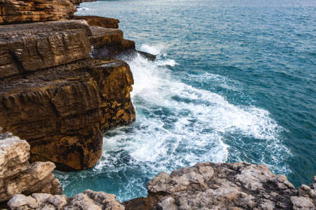 High brown layered rocks with ledges against which the waves of the turquoise ocean beat and break into white foam. scenic viewの写真素材
