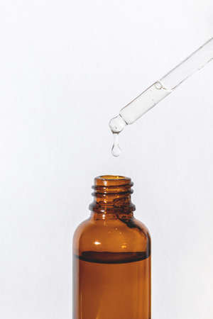 Dripping serum from a pipette with a vial on the side into a glass brown bottle on a white background, vertical close-upの写真素材
