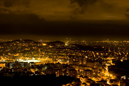 Night panorama of Barcelona. City lights, cloudy sky, and a dramatic urban landscape.の写真素材