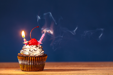 Birthday cupcake with blown out and burning candles and a red cherry on top over deep blue background in a cloud of a smokeの写真素材