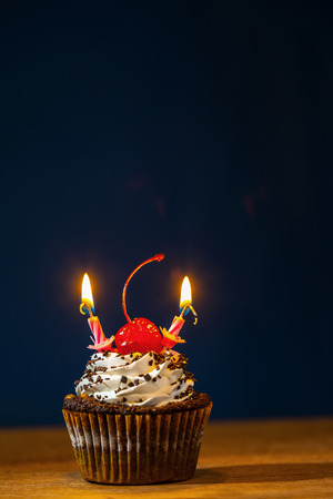 Birthday cupcake with red cherry and two burning candles on top over dark backgroundの写真素材