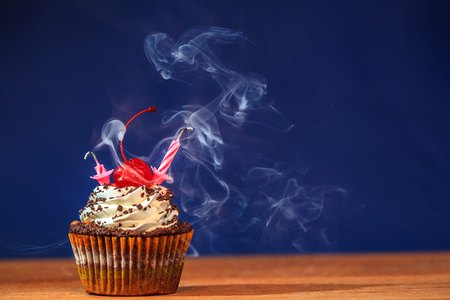 Birthday cupcake with two blown out candles and a red cherry on top over deep blue background in a cloud of a smokeの写真素材