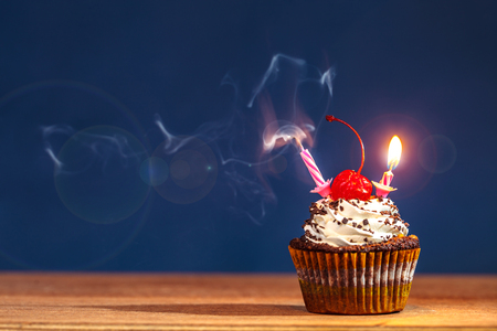 Birthday cupcake with blown out and burning candles and a red cherry on top over deep blue background in a cloud of a smoke. Lightflare filterの写真素材