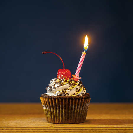 Birthday cupcake with burning candle and a red cherry on top over deep blue background の写真素材