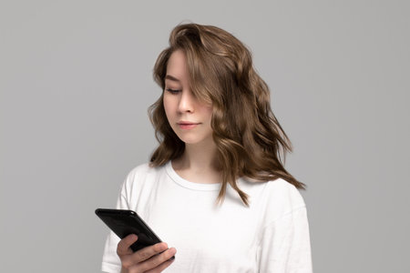 Portrait of a young woman dressed in white t-shirt using mobile phone, gray background, copy space....