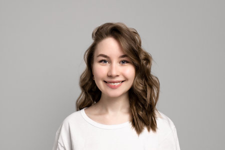 Portrait of happy smiling young woman looking at camera with natural everyday makeup, gray background, copy space. Beautiful caucasian girl with brown curly hairstyle. Cheerful expression faceの写真素材