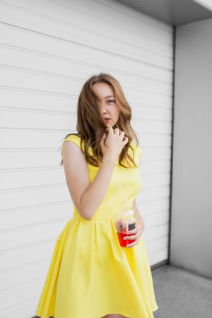 Young woman in illuminating yellow dress trend color of 2021 standing in front of white wall, holding glass of lemonade and looking at camera. Girl having fun. Urban style and fashion. Casual outfitの写真素材