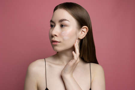 Young woman applying moisturising cream, lotion on face with fingers on pink background Healthy treatment and cosmetology products. Girl cleaning face. Beauty routine, skincare conceptの写真素材