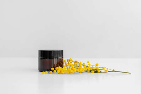 Cream ,serum, oil, lotion in brown glass jar and yellow mimosa flower, beige background. Zero waste...