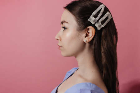 Face profile of young woman in blue shirt with natural makeup and curly hairstyle looking to the left over pink background, shadow. Girl in casual outfit in pearl earrings and hairpins studio shot.の写真素材