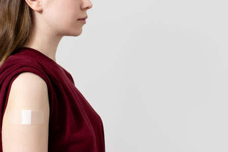 Unrecognizable woman in t-shirt showing plaster on shoulder after coronavirus vaccine, gray...