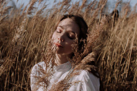 Young woman with closed eyes in white blouse sitting in field of pampas grass in front of sky and...