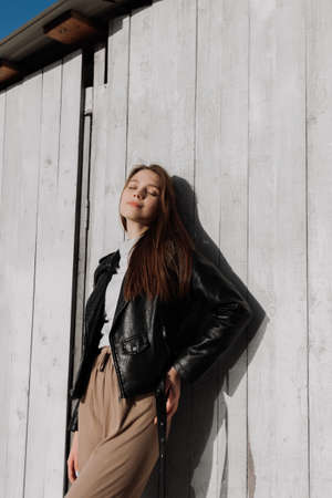 Smiling young woman in black leather jacket standing in front of white wooden wall. Having fun outside. Urban style and street fashion. Girl in casual outfit looking at cameraの写真素材