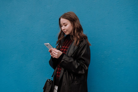 Smiling young woman in black leather jacket standing in front of blue wall and using smartphone. Urban style and street fashion. Girl looking down texting on smartphone. Chatting, messaging, readingの写真素材
