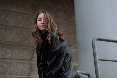 Smiling young woman in black leather jacket standing in front of gray wall, leaning on column, looking at camera. Urban style and street fashion. Girl with curly hairstyle in casual outfitの写真素材