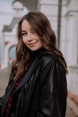 Young woman in black leather jacket and sweater standing in front of white wall looking at camera. Urban style and street fashion. Smiling girl with curly hairstyle in casual outfitの写真素材
