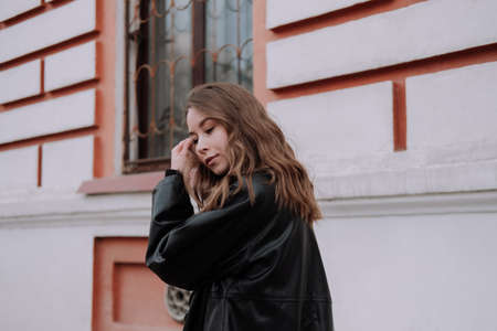 Smiling young woman in black leather jacket standing in front of white wall touching hair, looking...