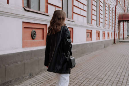 Unrecognizable woman in black leather jacket and white jeans walking on the street in front of white...