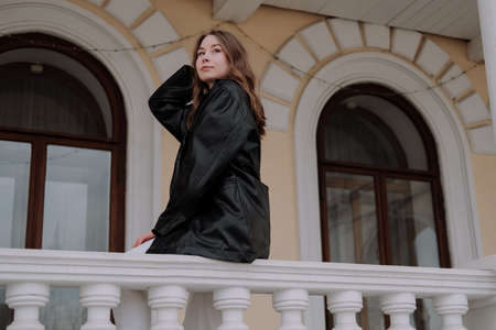 Smiling young woman in black leather jacket sitting on marble balustrade, balcony in front of yellow...
